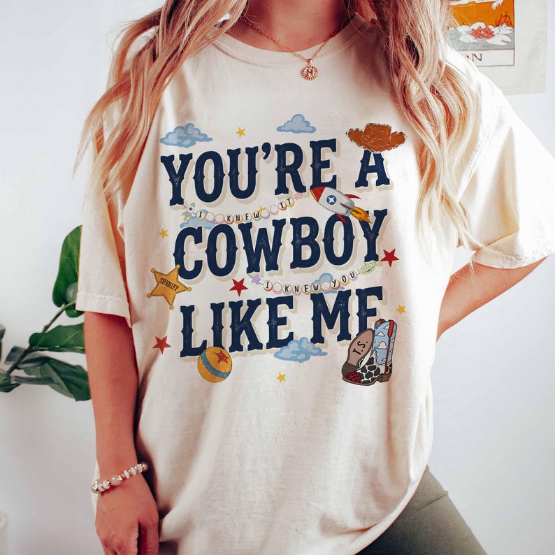May include: A cream-colored short-sleeve t-shirt featuring the dark blue text "YOU'RE A COWBOY LIKE ME" with smaller text "I KNEW I KNEW YOU". The graphic includes a rocket, a sheriff star, a ball, a cowboy hat, and a cowboy boot with "I.S.".