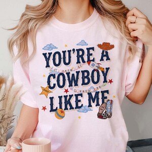 May include: A light pink t-shirt featuring the dark blue text "YOU'RE A COWBOY LIKE ME" with smaller "I KNEW IT I KNEW YOU" text on a beaded string. Graphics include a rocket, a sheriff star, a brown cowboy hat, a ball, and a patterned cowboy boot.