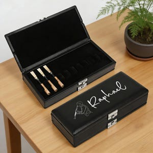 May include: Black leather clarinet reed case with silver clasps. The open case reveals reeds and storage slots. The closed case has the name "Raphael" and a clarinet player illustration in white.