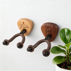 May include: Two wooden guitar wall hangers. One is light-colored with the name "Franklin" and the other is dark-colored with the name "James". Both have brown support arms and black rubber ends. The hangers are mounted on a white wall.