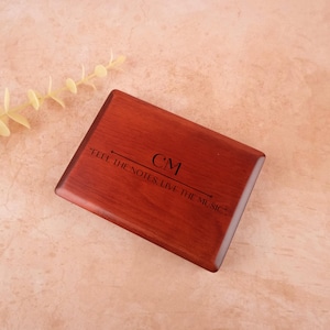 May include: A rectangular, wooden box with a rich, reddish-brown finish. The box features the initials "CM" and the phrase "FEEL THE NOTES, LIVE THE MUSIC" engraved on the surface. The box is likely a musical instrument case or a decorative item.
