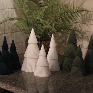 May include: A selection of decorative paper Christmas trees in different sizes and colours. The trees are arranged on a countertop, with some in dark green and others in white. A fern is in the background.