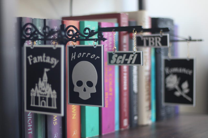 May include: Black metal shelf sign with hanging genre labels for books. Labels include "Fantasy" with a castle, "Horror" with a skull, "Sci-Fi", "TBR", and "Romance" with a rose. Books of various colors are in the background.