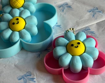 Peut inclure: Des boîtes à bijoux en forme de fleur, de couleur bleu clair et rose. Chaque boîte a un centre jaune avec un visage souriant et des pétales bleu clair. Les boîtes sont sur un fond de papier à motifs.