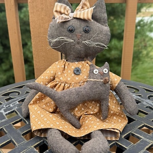 May include: A handcrafted fabric cat doll with dark brown textured skin, button eyes, and stitched whiskers, wearing a mustard yellow dress with a white floral pattern and a striped bow on its head, holding a smaller matching brown fabric cat.