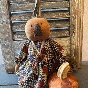 May include: A fabric pumpkin doll with a round orange head, a dark brown felt nose, and a stem on top. The doll wears a dress made of a multi-coloured patchwork fabric featuring floral and geometric patterns. A small metal jack-o'-lantern bell hangs from a ribbon around the neck, and a separate round fabric pumpkin sits at the base.