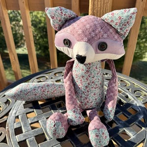 May include: A handmade stuffed fox doll with a floral pattern in shades of pink and green. The fox has button eyes, a black nose, and a long tail. The doll is sitting on a decorative metal table.