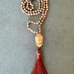 May include: A mala necklace featuring numerous round, light brown beads with dark brown veining. A carved, light-colored Buddha head pendant sits above a long, vibrant rust-orange silk tassel. The beads are strung in a continuous loop design.