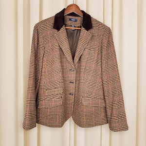 May include: A brown plaid blazer with a dark brown velvet collar, hanging on a wooden hanger. The blazer has a classic design with three buttons and two front pockets. The plaid pattern includes shades of brown, orange, and pink.