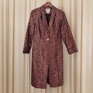 May include: A long, leopard-print coat in shades of brown and pink hangs on a wooden hanger. The coat features a single gold button closure and a notched collar. The lining is black. The label reads "KB".