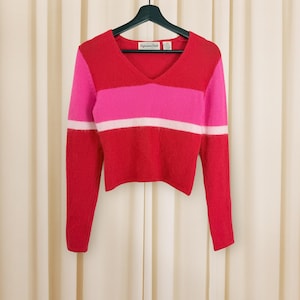 May include: A cropped, long-sleeve sweater in a color block design. The sweater features a V-neck and is primarily red, with a pink stripe across the chest and a white stripe below it. The sleeves are red with white stripes at the cuffs.
