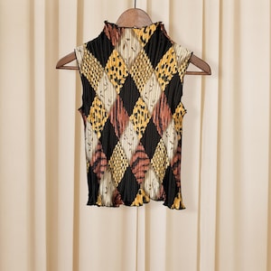 May include: A sleeveless top with a high ruffled collar, featuring a diamond pattern in black, yellow, and rust colors. The fabric is pleated, and the top hangs on a wooden hanger against a neutral background.