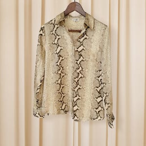 May include: A long-sleeved button-down shirt with a snakeskin pattern in shades of beige, brown, and black. The shirt is hanging on a wooden hanger against a neutral background. The collar is open, and the sleeves are cuffed.