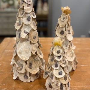 May include: Three decorative oyster shell Christmas trees of varying sizes. The trees are constructed from natural oyster shells, showcasing a range of off-white and tan hues with dark spots. The trees are arranged on a wooden surface.