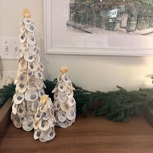 May include: Three decorative seashell Christmas trees of varying sizes. The trees are made from oyster shells and are arranged on a wooden surface. A framed picture of a building is in the background.