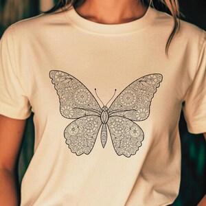May include: Ivory-colored t-shirt featuring a detailed black butterfly design. The butterfly's wings are filled with intricate patterns. The shirt is short-sleeved and has a round neckline. The word "ivory" is displayed at the bottom of the image.