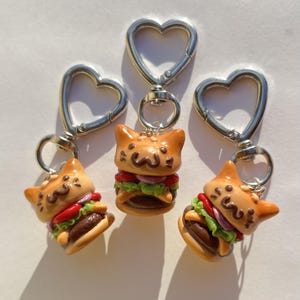 Könnte beinhalten: Drei neuartige Schlüsselanhänger mit einem katzenförmigen Burger-Design. Die Burger haben rote, grüne und violette Schichten. Jeder Schlüsselanhänger hat einen silbernen herzförmigen Verschluss.