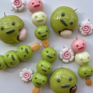 May include: A collection of handmade polymer clay charms featuring green, pink, and white dango, a Japanese sweet treat. The charms have smiling faces and are on skewers or shaped like flowers. Some charms have a metal loop for attaching to jewelry.