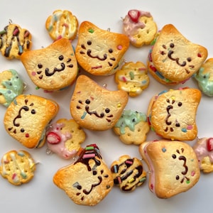 Puede incluir: Una colección de colgantes de comida en miniatura hechos a mano. Los colgantes incluyen sándwiches con forma de gato, galletas y crackers, decorados con glaseado colorido, chispas y salsa de chocolate. Los colgantes son de varias formas y tamaños, con una combinación de colores marrón claro y naranja.