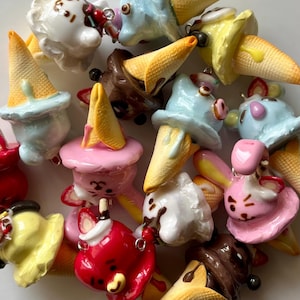 May include: A collection of colorful, miniature ice cream cone charms. The charms feature various colors of ice cream, including pink, blue, white, and brown, topped with a waffle cone. Some have small, decorative details.
