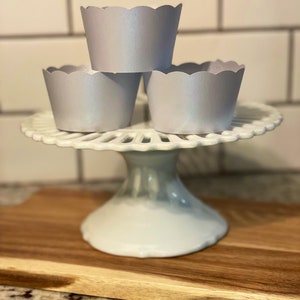 May include: Three silver cupcake liners with scalloped edges are stacked on a white cake stand. The stand is on a wooden surface.