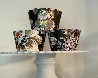 May include: Three black cupcake liners with a floral pattern in gold and white. The liners are stacked on a white cake stand.