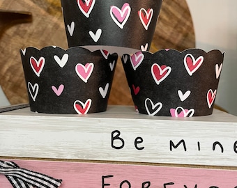 May include: Three black cupcake liners with a scalloped edge, decorated with pink, red, and white hearts. The liners are stacked on top of wooden blocks that read "Be mine FOREVER Lots of Love."