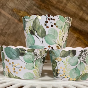 May include: Three white cupcake liners with a green and gold watercolor leaf pattern. The liners have a scalloped edge.