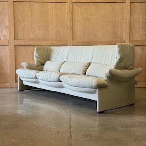 May include: A light beige sofa with three seat cushions and three smaller back cushions. The sofa has high, curved armrests and a tall back. The sofa is set against a light wood paneled wall and a concrete floor.