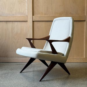 Peut inclure: Fauteuil de style milieu du siècle avec assise et dossier rembourrés de couleur crème clair. Le fauteuil présente des accoudoirs et des pieds en bois brun foncé. Le design est minimaliste, sur fond de panneaux de bois clair.