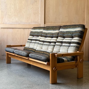 May include: A mid-century style sofa with a light wood frame and a striped cushion in shades of brown, beige, and black. The sofa has a low profile and a simple, geometric design, suitable for a living room or lounge area.