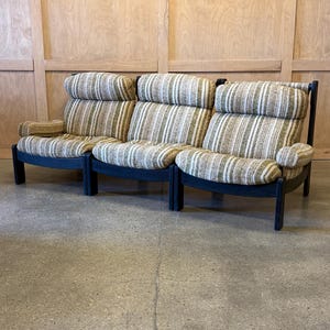 May include: A three-seat sofa with a high back and armrests. The sofa features a striped pattern in shades of beige, brown, and green. The frame is dark, and the sofa is set against a light wood panel wall.