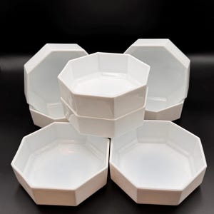 May include: A set of five white ceramic bowls, each featuring a distinct octagonal shape with smooth, angled sides and a flat base. The bowls are arranged in a cluster, with one bowl stacked directly on top of another in the center, surrounded by three additional bowls.