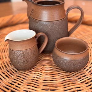 May include: A brown ceramic tea set with a textured finish. The set includes a teapot with a handle and spout, a creamer, and a small bowl. The interior of the creamer is white. The set is displayed on a woven wicker tray.
