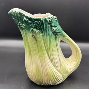 May include: A ceramic pitcher shaped like a head of lettuce or cabbage, featuring a textured surface with vertical ridges. The exterior displays a gradient of colors, transitioning from a deep forest green at the top rim to a pale cream at the base.