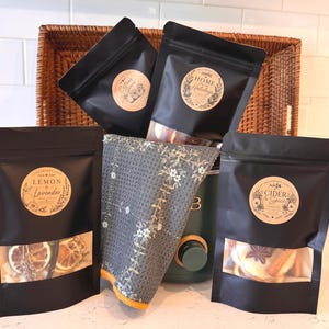 May include: Four black stand-up pouches with clear windows containing dried fruit and spices, labeled Lemon & Lavender, Citrus Grove, Home for the Holidays, and Cider & Spice. A grey waffle-weave towel with a white floral print rests in front.