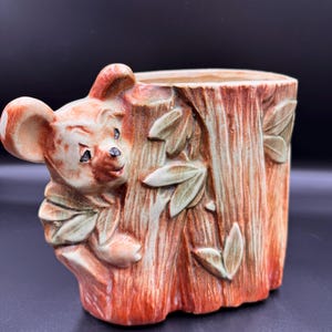 May include: A ceramic planter shaped like a tree stump with a cartoon mouse peeking out. The planter is primarily orange and brown with green leaves. The mouse has tan ears and a black nose. A decorative item.
