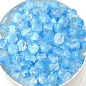 Puede incluir: Una colección de cuentas de vidrio azul claro con un diseño arremolinado y texturizado. Las cuentas son redondas y varían ligeramente en forma, con una calidad translúcida. Están contenidas en un cuenco blanco, creando un contraste con el azul.