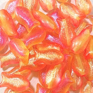 以下が含まれることがあります： オレンジとピンクのイリデセントな魚の形をしたビーズのコレクション。ビーズは光沢のある仕上がりで、サイズと形が異なり、星に似たものもあります。ビーズは白い表面に散らばっています。