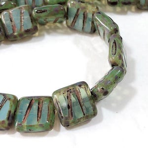May include: A strand of square glass beads featuring a translucent light blue base with a glossy green and brown mottled finish. Each bead displays three parallel dark brown etched lines across the surface, creating a textured appearance.