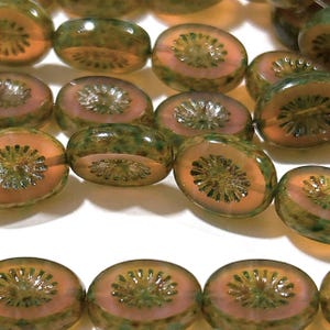 May include: Close-up of several glass beads with a kiwi fruit design. The beads have a translucent appearance, with a light orange center, a pinkish-red ring, and a green outer layer. The beads are oval-shaped and appear to be handmade.