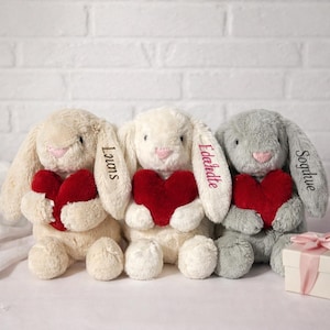Puede incluir: Tres conejos de peluche en crema, blanco y gris, cada uno sosteniendo un corazón rojo. Los conejos tienen nombres bordados en sus orejas. Una pequeña caja de regalo con una cinta rosa está a la derecha.