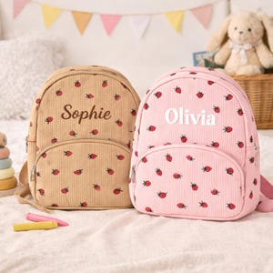 May include: Two corduroy backpacks, one beige and one pink, are decorated with red strawberry patterns. The beige backpack has the name "Sophie" embroidered on it, while the pink backpack has "Olivia". Both have a front pocket and adjustable straps.