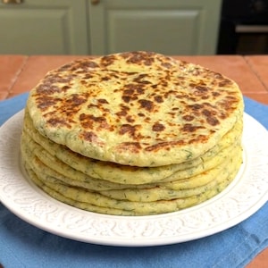 May include: A stack of golden-brown, round flatbreads on a white plate. The flatbreads have a slightly speckled appearance with visible green herbs. The plate sits on a blue cloth, with a wooden table and green background.