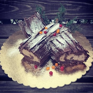 May include: A chocolate Yule log cake, cut to reveal a creamy filling, sits on a gold scalloped cake board. The cake is decorated with chocolate bark, icing sugar, and colourful sweets. Sprigs of greenery add a festive touch.