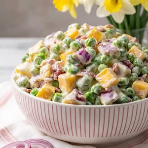 May include: A bowl of pea salad with diced cheddar cheese, red onion, and bacon. The salad is mixed with a creamy dressing and served in a pink and white striped bowl. Yellow daffodils are in a vase in the background.
