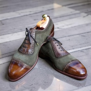 Könnte beinhalten: Ein Paar Oxford-Schuhe aus einer Kombination von olivgrünem Wildleder und poliertem braunem Leder. Die Schuhe haben eine Zehenkappe mit dekorativen Brogue-Details entlang der Nähte, dunkelbraune Schnürsenkel und einen hölzernen Schuhspanner.