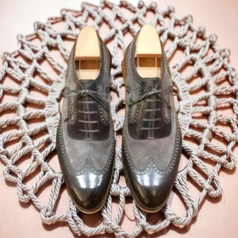 Puede incluir: Un par de zapatos de vestir de cuero gris y negro con hormas de madera. Los zapatos tienen un dise&ntilde;o de punta de ala y est&aacute;n atados con cordones. Se exhiben sobre una estera circular tejida.