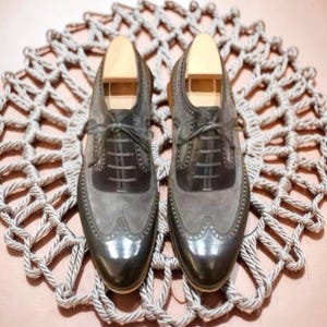 Puede incluir: Un par de zapatos de vestir de cuero gris y negro con hormas de madera. Los zapatos tienen un dise&ntilde;o de punta de ala y est&aacute;n atados con cordones. Se exhiben sobre una estera circular tejida.