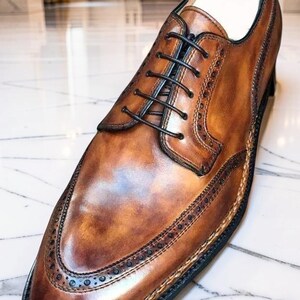 May include: A close-up of a brown leather dress shoe with black laces. The shoe features decorative stitching and a wingtip design. The shoe is displayed on a marble surface.